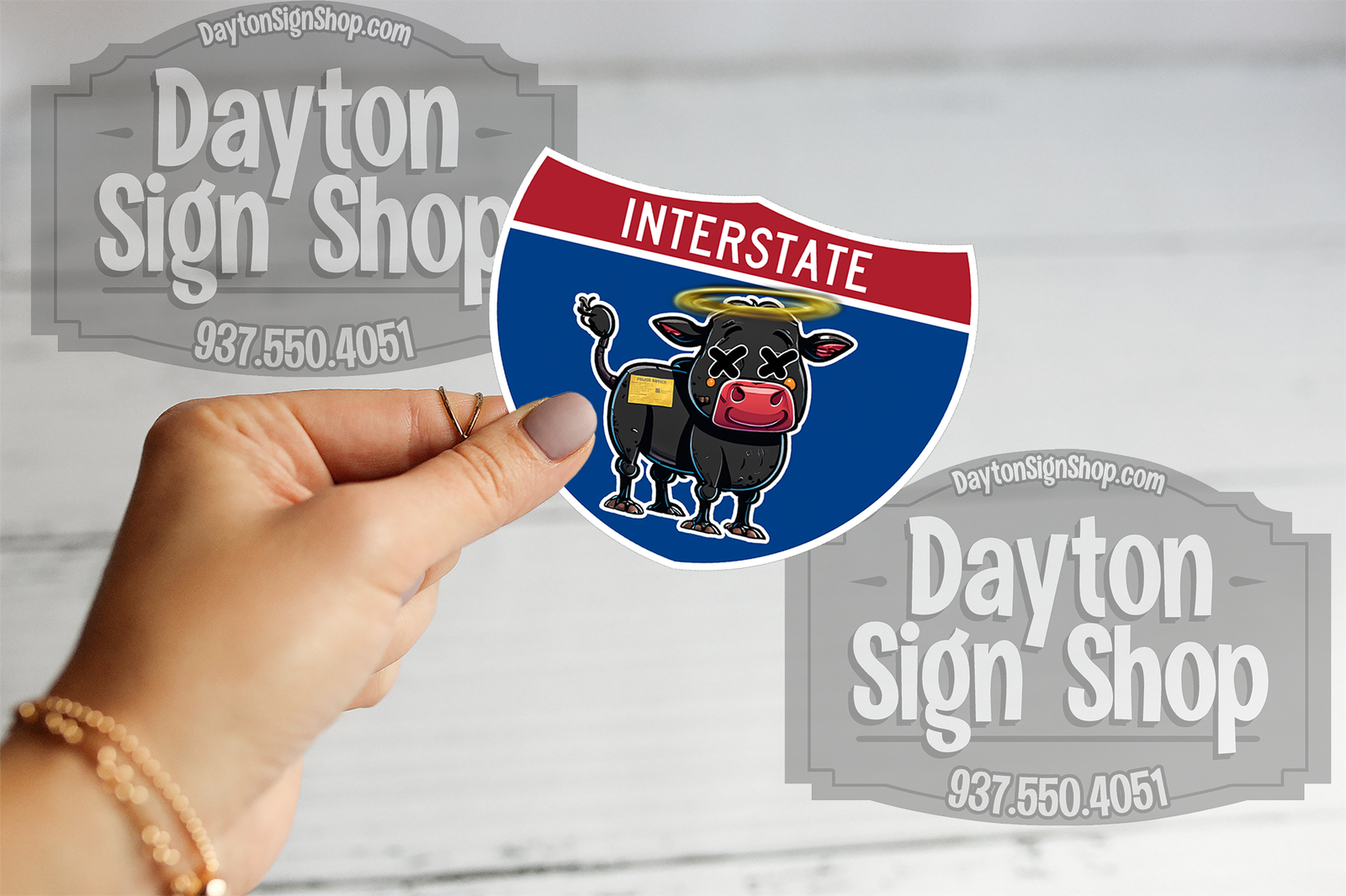 Custom cow sticker held in front of Dayton Sign Shop stickers